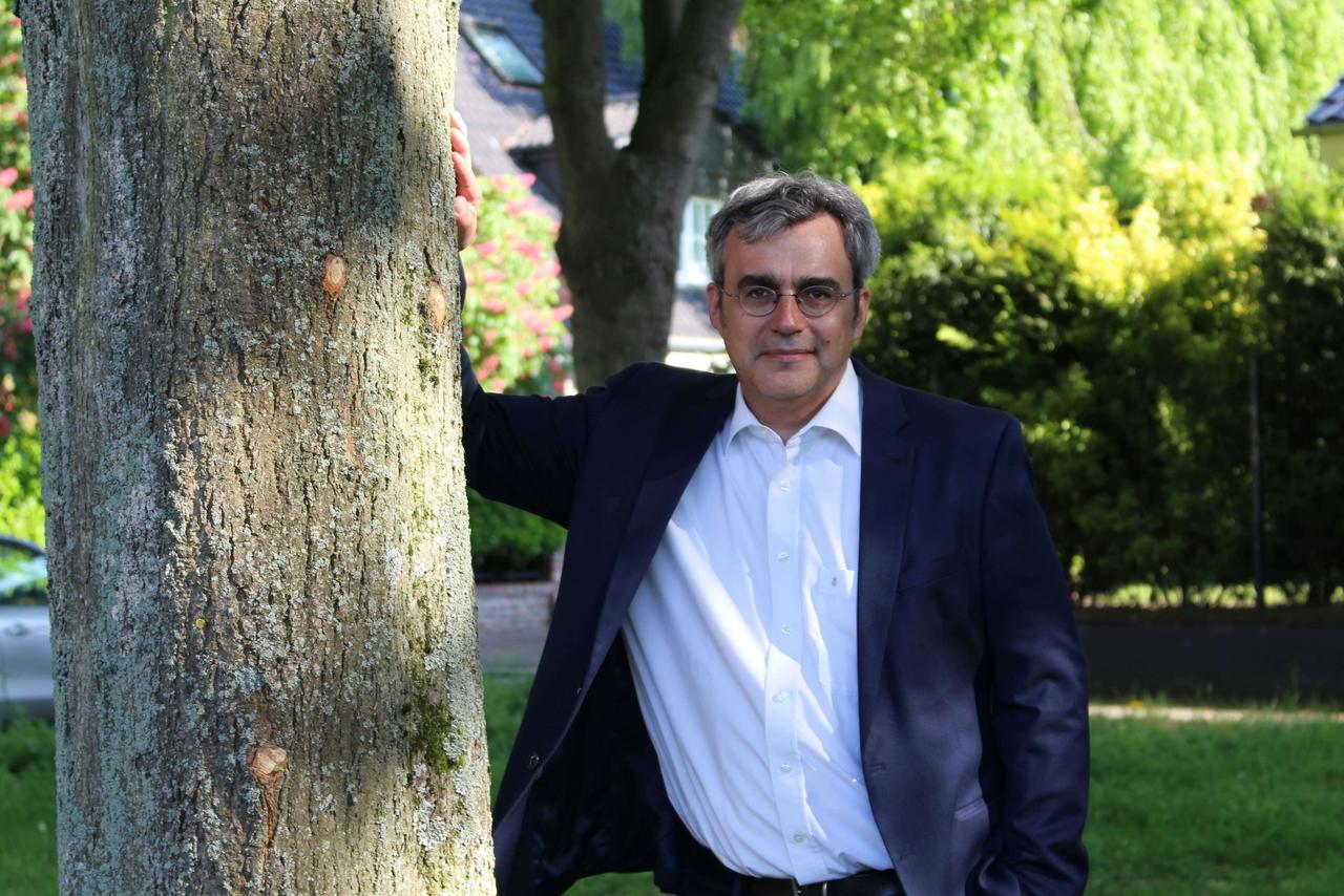 Ein Mann lehnt sich draußen an einen Baum, macht kurz Pause. Im Hintergrund sind Büsche und ein Gebäude zu sehen.