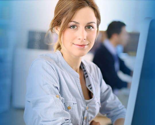 Eine Frau arbeitet am Schreibtisch, schaut in die Kamera. Im Hintergrund ist ein Kollege bei seiner Arbeit zu sehen.