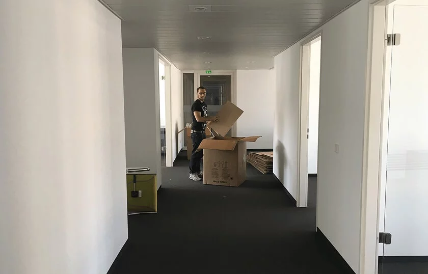 Jemand faltet Umzugskartons in Büroflur, nebenan stapeln sich leere Boxen und Verpackungsmaterial.